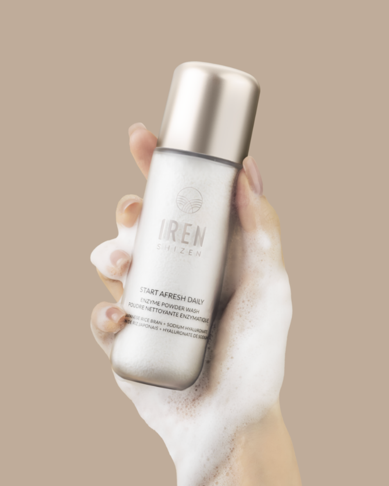 A hand covered in foam holds a bottle of IRÉN Shizen START AFRESH DAILY Enzyme Powder Wash, a gentle rice enzyme cleanser and daily exfoliator, against a plain beige background.