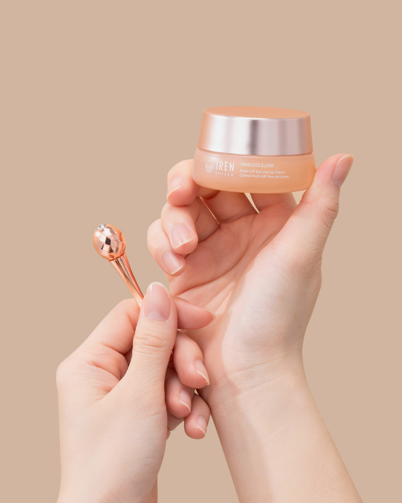 A person holds IREN Shizen's TIMELESS ELIXIR Multi-Lift Eye and Lip Cream in one hand and a rose gold applicator in the other, showcasing the anti-aging formula against a plain beige background.