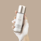 A hand covered in foam holds a bottle of IRÉN Shizen START AFRESH DAILY Enzyme Powder Wash, a gentle rice enzyme cleanser and daily exfoliator, against a plain beige background.