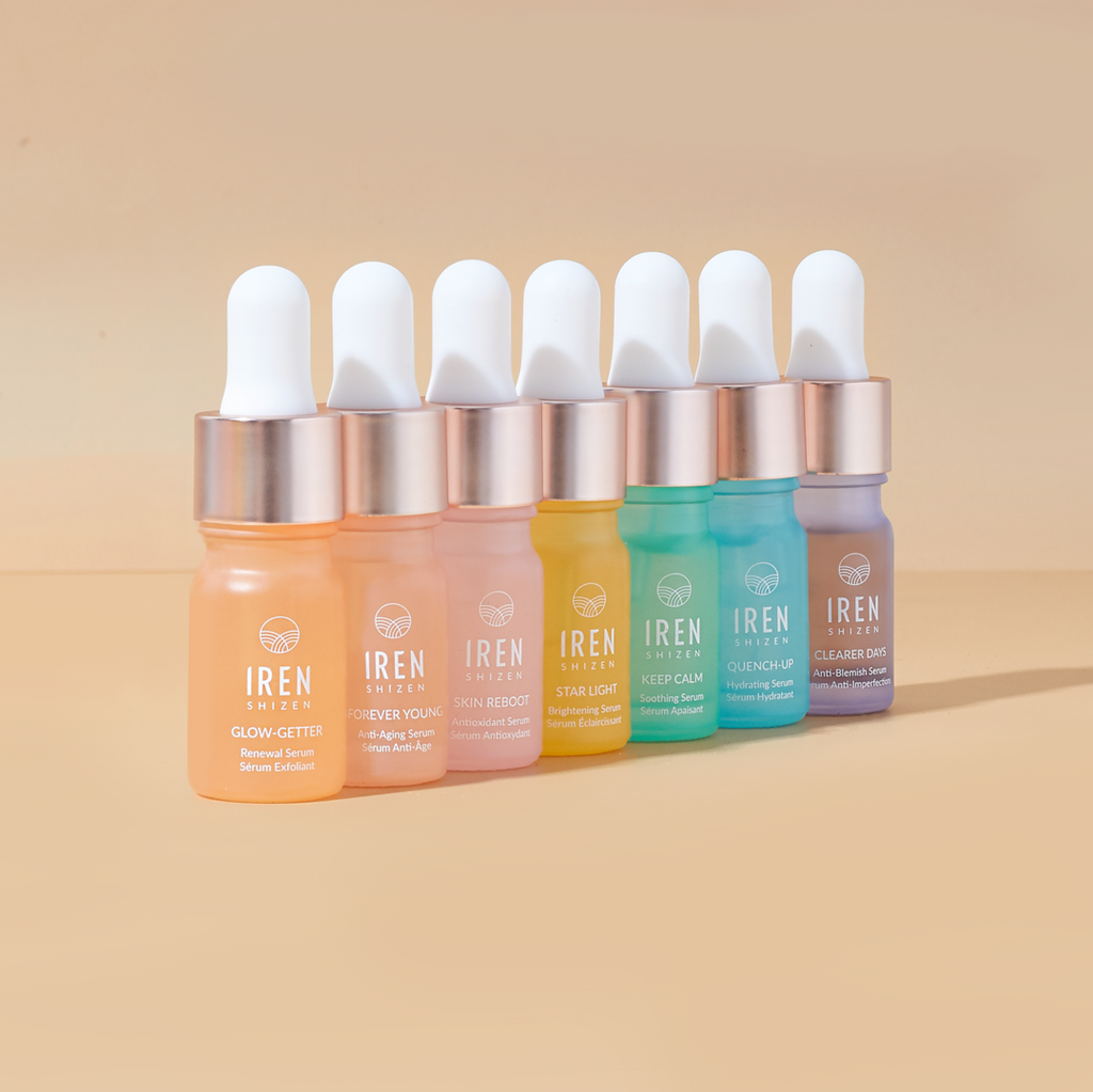 Seven colorful bottles of MOCHI SKIN Instant Glow Travel Set skincare serums from the IREN Shizen lined up against a peach-colored background.