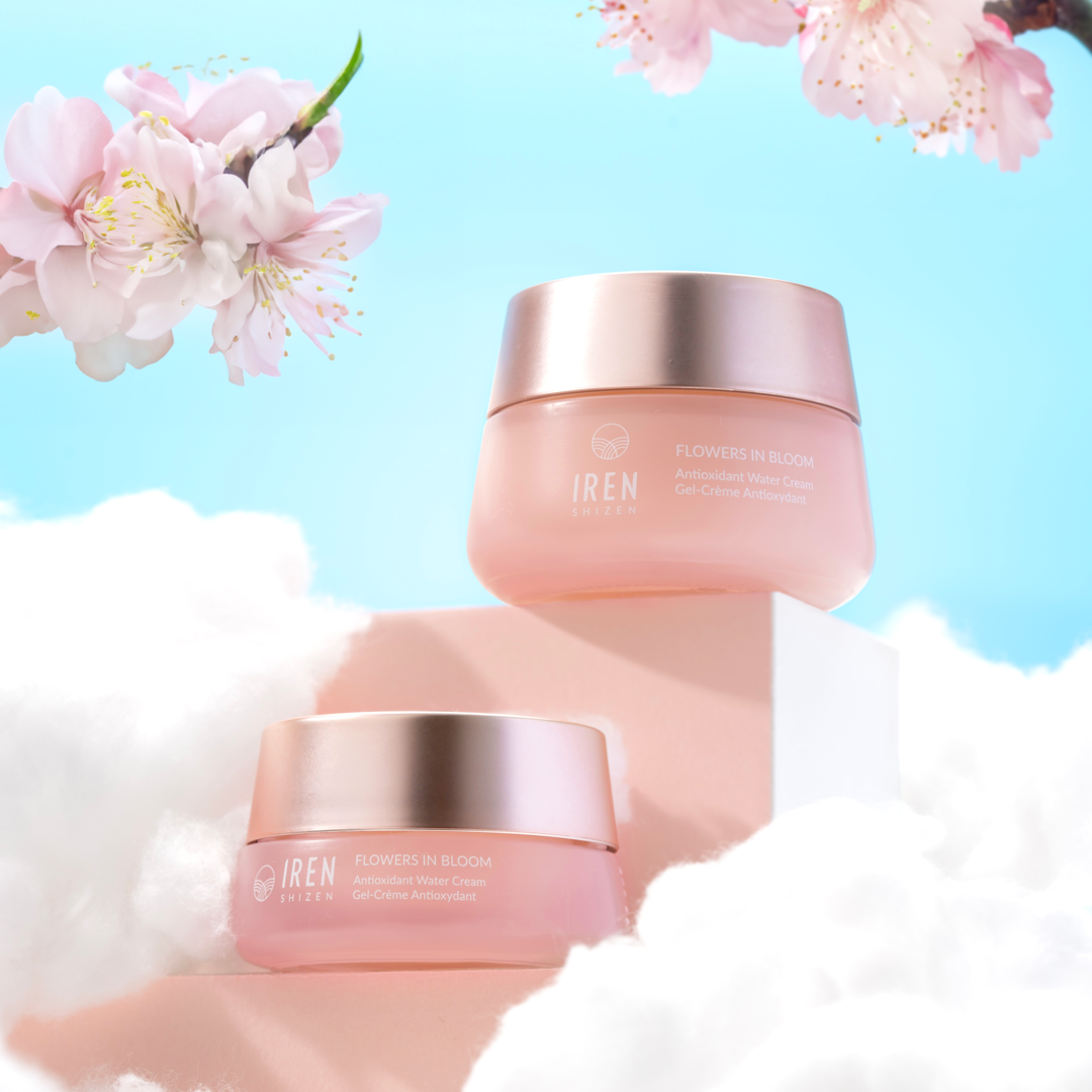 Two jars of "FLOWERS IN BLOOM" Antioxidant Water Cream by IREN Shizen are gracefully showcased on a cloud-like surface, surrounded by blooming cherry blossom branches and set against a clear blue sky. This silicone-free gel cream is ideal for sensitive skin, offering hydration without irritation.