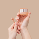 A person holds IREN Shizen's TIMELESS ELIXIR Multi-Lift Eye and Lip Cream in one hand and a rose gold applicator in the other, showcasing the anti-aging formula against a plain beige background.