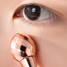 Close-up of an eye with a rose gold-colored facial massager rolling under the lower eyelid, promoting anti-aging and rejuvenation. Featured alongside is IREN Shizen's TIMELESS ELIXIR Multi-Lift Eye and Lip Cream.