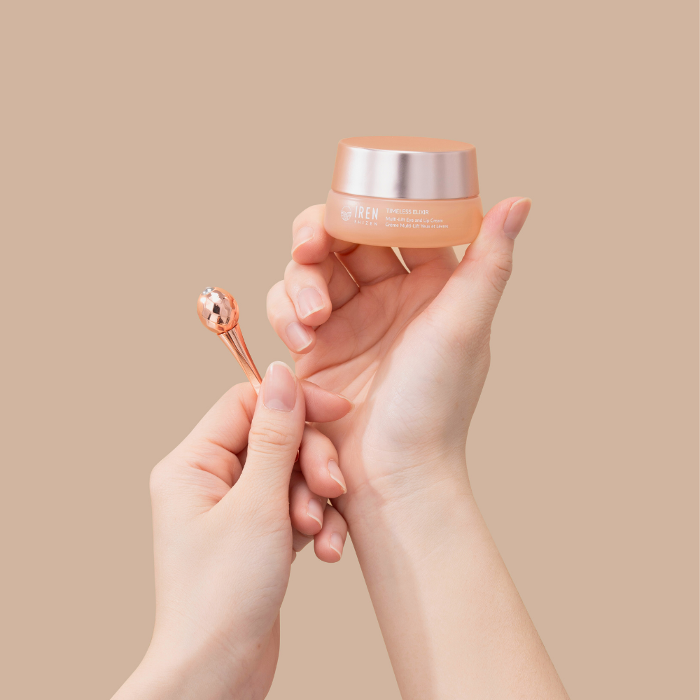 A person holds IREN Shizen's TIMELESS ELIXIR Multi-Lift Eye and Lip Cream in one hand and a small applicator with a metallic ball in the other, poised to target dark circles against a beige background.