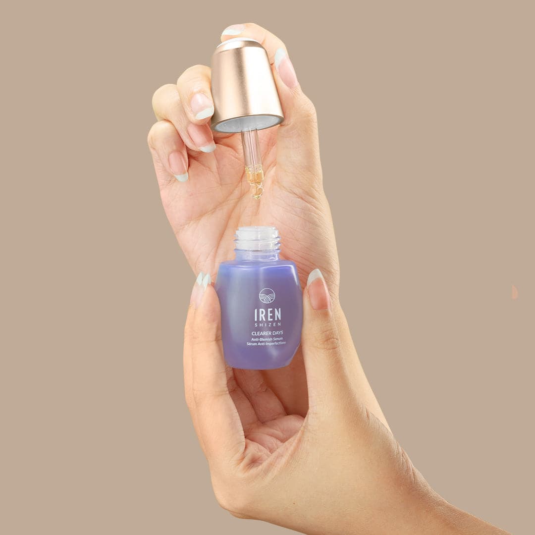 A hand holds a blue glass dropper bottle labeled "IREN Shizen," identifying it as the CLEARER DAYS Anti-Blemish Serum. The dropper, lifted from the bottle, drips clear liquid—a potent solution—against a plain beige background.