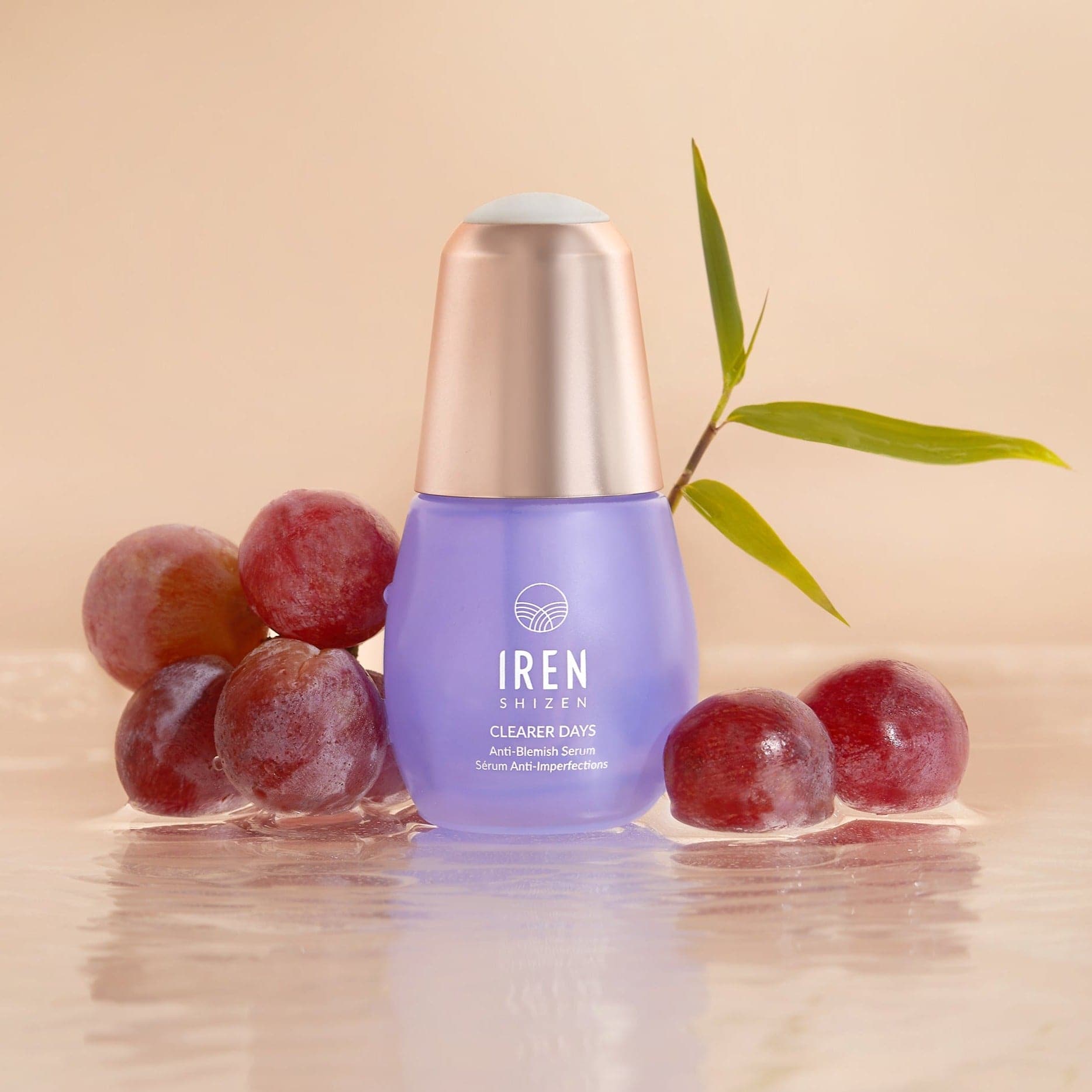 A purple bottle of CLEARER DAYS Anti-Blemish Serum from IREN Shizen is nestled among red grapes and a bamboo leaf on a reflective surface, showcasing its prowess as an anti-blemish solution.